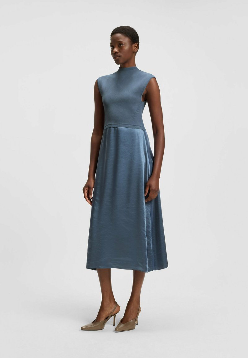 A sleeveless, midi-length dress in a soft, muted blue fabric with a fitted bodice and flared skirt, paired with neutral high-heeled shoes.
