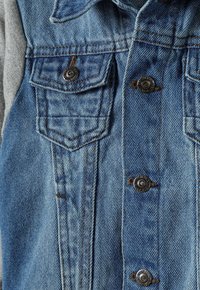 Chaqueta de denim con un lavado azul claro, que presenta dos bolsillos en el pecho, herrajes de bronce y acentos de tela gris en las mangas. Acabado texturizado.
