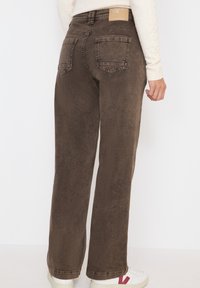 Pantaloni in denim marrone con design a gamba dritta, tasche posteriori e un'etichetta in pelle beige in vita. Abbinati a sneakers bianche.