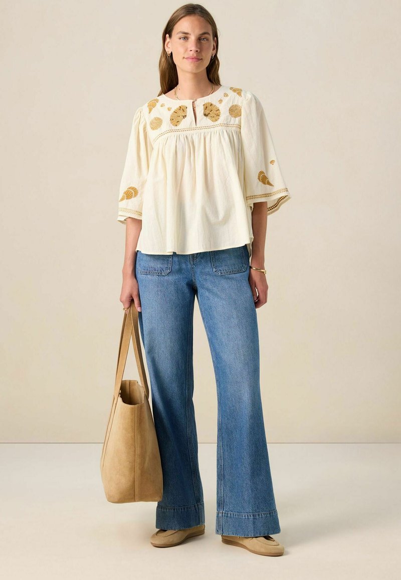Weiße, bestickte Bluse mit goldenen Akzenten, kombiniert mit weit geschnittenen blauen Jeans. Das Model trägt eine hellbraune Stofftasche. Flache Schuhe runden den Look ab.