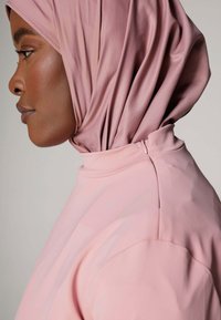 Lichtroze hijab van zacht, glad materiaal, elegant gedrapeerd. De outfit heeft een hoge hals met een discrete ritssluiting aan de achterkant.