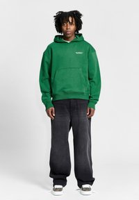 Groene katoenen hoodie met een voorzak, voorzien van ribbelboorden. Gecombineerd met losse zwarte denim broeken en grijze sneakers met witte accenten.