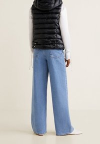 Schwarze Pufferweste mit Kapuze, kombiniert mit hellblauen weiten Jeans. Die Jeans haben eine klassische Denim-Textur und sichtbare Nähte.