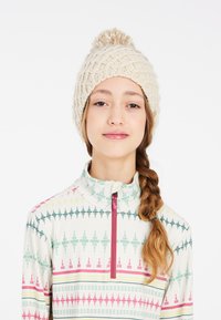 Bonnet en tricot crème avec un pompon sur le dessus. Le modèle porte un haut zippé à motifs dans des couleurs douces avec des designs géométriques.