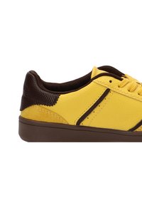 Zapatilla amarilla con cuero texturizado y gamuza, acentos marrones, suela marrón lisa y sistema de cordones tradicional.