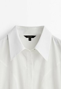 Chemise à col blanc en tissu lisse, présentant une fermeture à boutons, des manches courtes et un léger brillant, avec une étiquette indiquant la taille et l'origine.