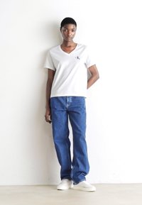 Tricou alb cu decolteu în V și logo mic, blugi din denim albastri și adidași albi, stând în fața unui fundal alb. Design simplu, îmbrăcăminte casual.