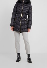 Schwarze Steppjacke mit Kapuze und Kunstpelzbesatz, ausgestattet mit einem Taillengürtel, goldenen Reißverschlüssen und einem Chevron-Steppdesign.