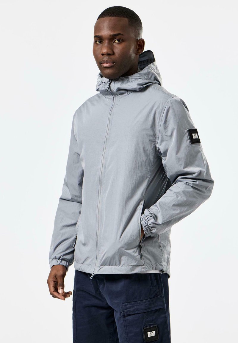 Light gray zip-up jacket with a hood, textured fabric, elastic cuffs, and a black patch on the left sleeve. Side pockets with zippers.