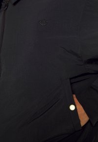 Černá lehká bunda s texturovaným povrchem, disponující zipovým zapínáním, malým vyšívaným logem a detailním patentním knoflíkem na kapse.