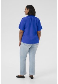 Blouse courte à manches courtes en tissu bleu texturé avec un col froncé, associée à un jean droit bleu clair et des ballerines noires. Vue arrière montrée.