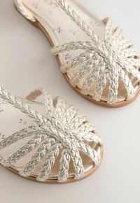 Flat sandals with braided silver straps forming a starburst pattern over a metallic insole. The sole is tan with a floral print.