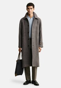 Homme portant un long manteau en pied de poule marron, une chemise bleu clair, un pantalon marron, des chaussures noires, tenant un sac fourre-tout noir avec des poignées marron.