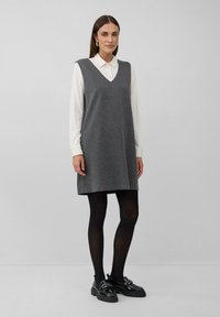 Vrouw in een witte lange mouwen shirt onder een grijze mouwloze V-hals jurk, zwarte panty en zwarte instappers, staand tegen een effen achtergrond.