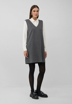Frau in einem weißen Langarmshirt unter einem grauen ärmellosen V-Ausschnitt-Kleid, mit schwarzen Strumpfhosen und schwarzen Slippern, vor einem einfarbigen Hintergrund stehend.