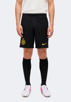 Czarne sportowe spodenki z żółtym logo Interu Mediolan i znakiem Nike, noszone z czarnymi skarpetami i białymi korkami na osobie stojącej nieruchomo.
