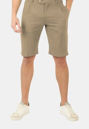 Uomo che indossa pantaloncini beige fino al ginocchio e scarpe da ginnastica bianche, in piedi con le mani parzialmente nelle tasche.