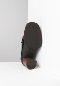Chaussure en cuir noir avec un talon en bois brun. La semelle présente une texture nervurée et un marquage. Le talon est de forme carrée et robuste.