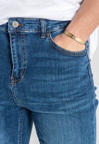 Blue denim jeans with a high waist, front pocket, and contrast stitching. Features a silver button and a gold bracelet on the wrist.