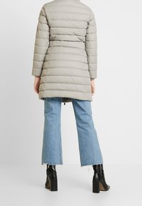 Gevoerde, lichtgrijze jas met een cinched taille, lange mouwen en een geborduurd ontwerp; gecombineerd met blauwe wijde jeans en zwarte enkellaarzen.