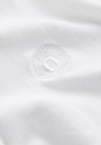 Biała tkanina z wypukłym haftowanym logo otoczonym wieńcem laurowym, charakteryzująca się gładką teksturą i klasycznym wzornictwem.