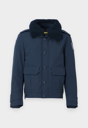 Navy blue jacket with a fluffy, dark collar, two front pockets, button closure, and logo patch on the sleeve. Structured shape and smooth texture.