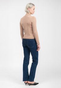 Mujer con cabello rubio corto, vistiendo un suéter de mangas largas beige, jeans azul oscuro y zapatos de tacón bajo negros, de pie de perfil sobre un fondo blanco.