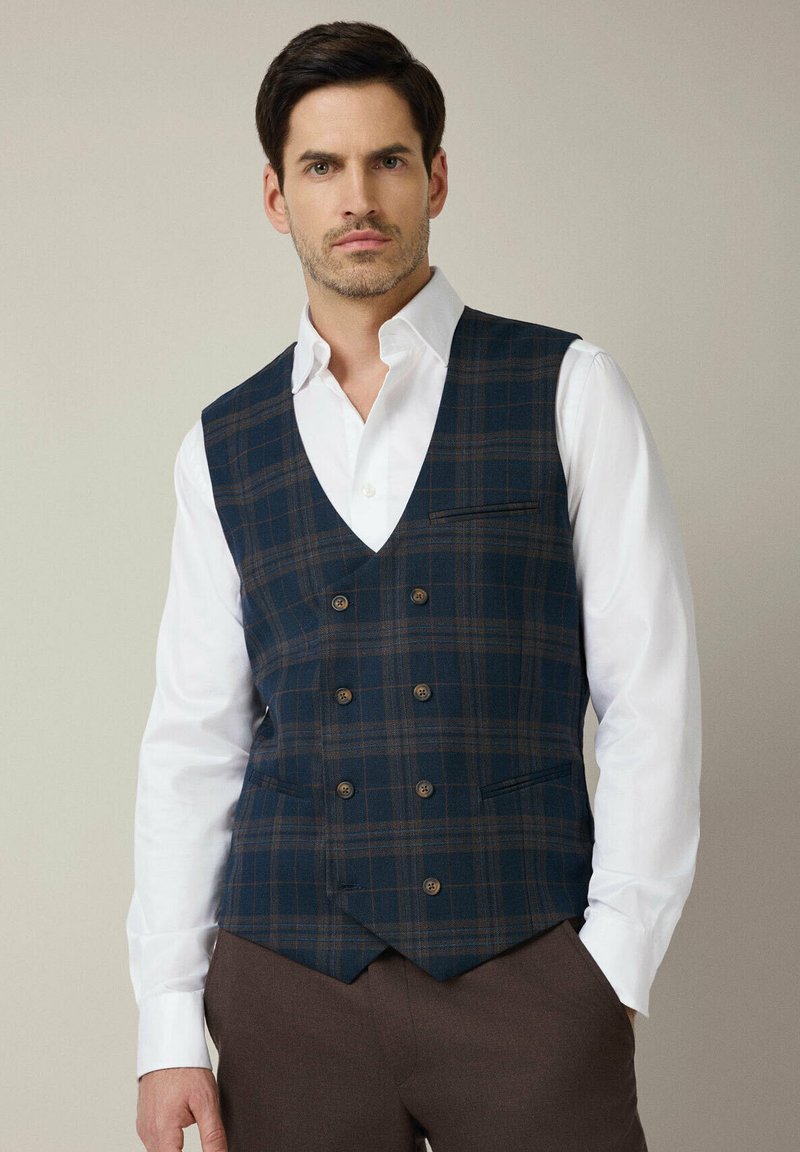Ciemnoniebieska kamizelka w kratę o dopasowanym kroju, z dekoltem w serek, podwójnym zapięciem na guziki i małymi bocznymi kieszeniami, noszona na białej koszuli.