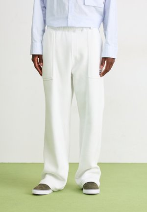 Mężczyzna w luźnych białych spodniach z przednimi kieszeniami naszywanymi i szarych butach wsuwanych, stojący na jasniziołtym podłożu.