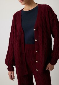 Persona valkā bordo krāsas adīto džemperi ar sudraba pogām, saskaņotās biksēs un tumši zilā t-kreklā uz neitrāla fona.