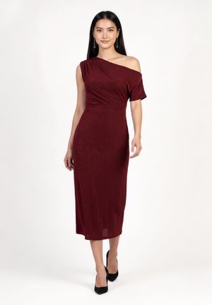 Femme portant une robe midi bordeaux ajustée à une épaule et des talons noirs, debout devant un fond blanc uni.