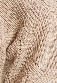 Pull en maille beige avec un motif diagonal texturé, doté de petites ouvertures le long des coutures pour un détail supplémentaire.