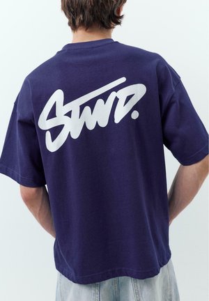 Férfi sötétkék pólóban, amelynek felső hátulján nagy, stilizált fehér "SWD." felirat látható, világos háttér előtt áll.