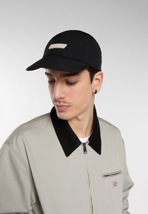 Nuori mies, jolla on musta lippalakki ja beige takki mustalla kauluksella, katsoo alas ja sivulle yksiväristä taustaa vasten.