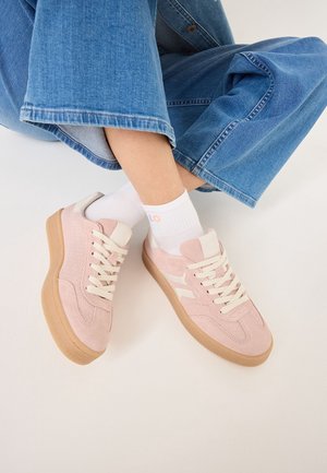 Person iført lyserøde ruskindssneakers med hvide snørebånd og gummisåler, hvide sokker og blå vidde jeans siddende mod en hvid baggrund.
