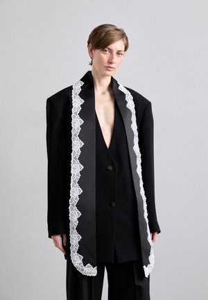 Blazer nero con una silhouette oversize, caratterizzato da un bordo in pizzo bianco lungo la parte anteriore, bottoni su misura e una profonda scollatura a V.