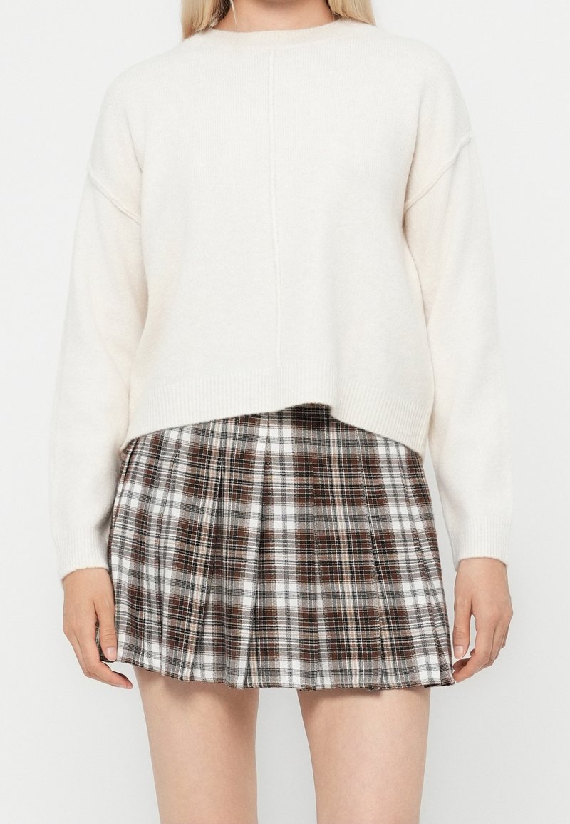 Femme portant un pull blanc à manches longues et une jupe plissée à carreaux marron et blanc, debout devant un fond uni.