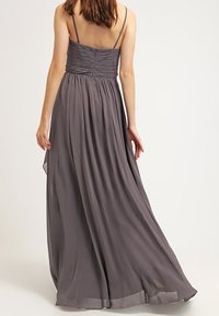 Žena viděná zezadu v dlouhých šedých šatech bez rukávů se tenkými ramínky a pletenou korzetovou částí.