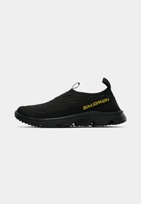 Black slip-on shoes with textured fabric, sporty design, and rubber sole. Features a yellow "SALOMON" logo on the side.