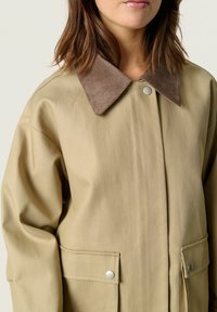 Veste d'extérieur beige avec un col en velours côtelé, poches avant fermées par des boutons-pression, présentant une texture lisse et mate.