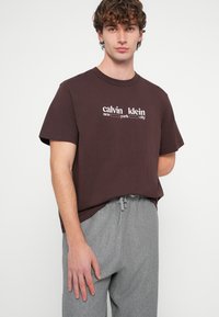 Rudas medvilninis marškinėlis su baltu prekės ženklo tekstu. Derinamas su pilkomis kelnėmis su raišteliais. Paprastas apvalus kaklas, laisvas siluetas.