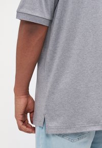 Camisetta grigia a maniche corte con spacchi laterali, tessuto testurizzato e vestibilità rilassata, abbinata a jeans in denim azzurro chiaro.