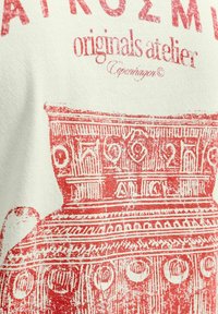 Weißer Stoff mit einem roten Grafikdruck eines alten Gefäßdesigns, das lineare Muster, kreisförmige Akzente und eine abgenutzte Textur aufweist.