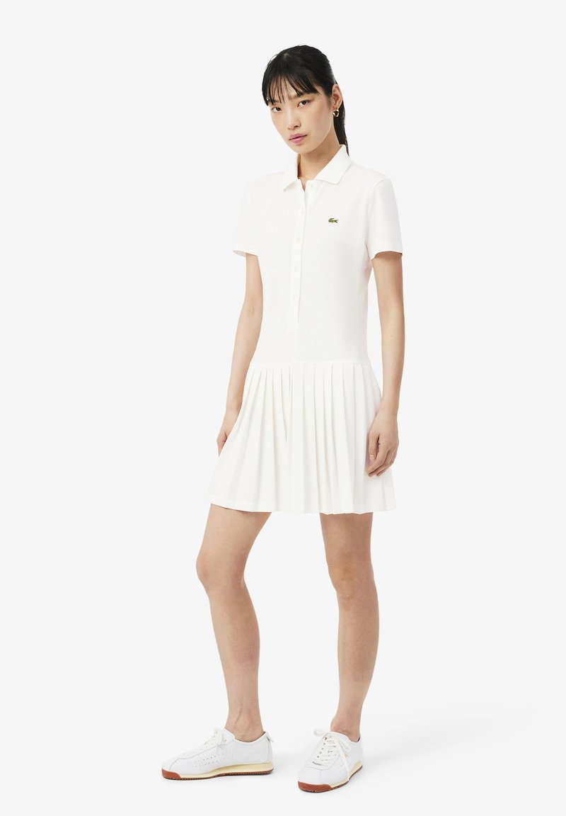 Jeune femme portant une robe polo blanche à manches courtes avec jupe plissée et baskets blanches, debout devant un fond blanc uni.