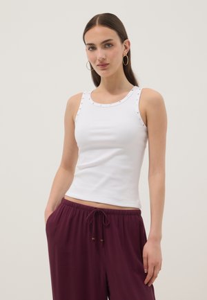 Vrouw met lang bruin haar die gouden oorringen draagt, een wit met studs versierd mouwloos topje en losse bordeauxroodgekleurde broek met trekkoord.