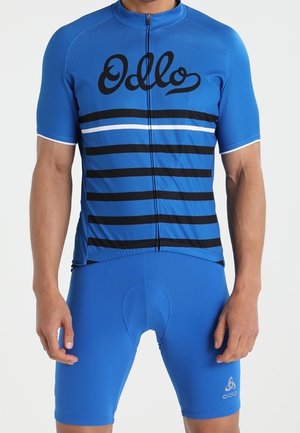Man draagt een blauw fietsjersey met zwarte strepen en bijpassende blauwe fietsshort, staand tegen een effen achtergrond.