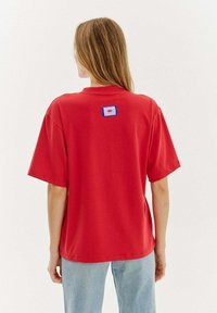 Czerwona bawełniana koszulka z krótkim rękawem, z niebieską i białą naszywaną łatką w kształcie oka z tyłu. Luźny krój z okrągłym dekoltem.