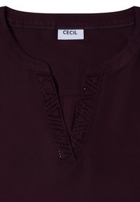 Bordeauxrotes Oberteil mit V-Ausschnitt, verziert mit dekorativen Perlen in geometrischem Muster entlang des Ausschnitts. Glatter Stoff mit matter Oberfläche.