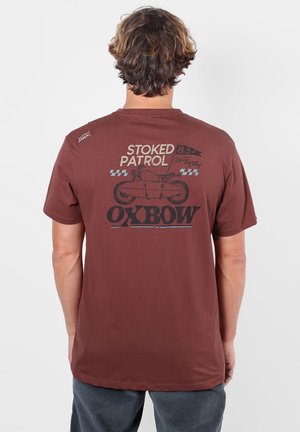 Homme aux cheveux ondulés portant un T-shirt marron avec "STOKED PATROL" et un motif de moto, debout devant un fond clair uni.
