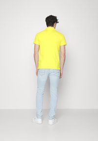 Sárga póló, rövid ujjú, sima anyagú. Világoskék, slim-fit farmerrel és fehér tornacipővel párosítva, hátulról látható.
